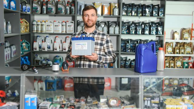 Smiling man in a plaid shirt holding a battery box behind the counter of an auto parts store with shelves of motor oil