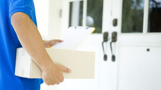 Delivery person in a blue shirt holding a cardboard package and a clipboard at a white front door