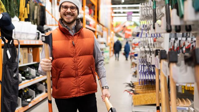 Smiling man in an orange vest and knit cap carrying an axe through a hardware store aisle