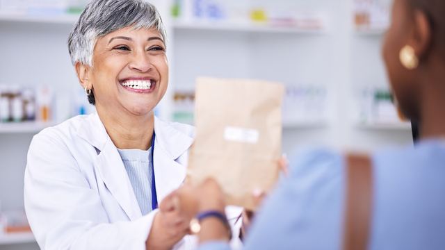 Smiling pharmacist in a white coat handing a paper pharmacy bag to a customer across the counter