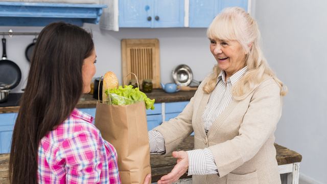 Young woman in a plaid shirt handing a paper bag of fresh vegetables to a smiling older woman in a kitchen