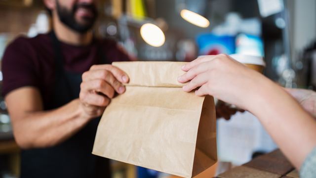 Man in an apron handing a brown paper takeout bag across a restaurant counter to a customer